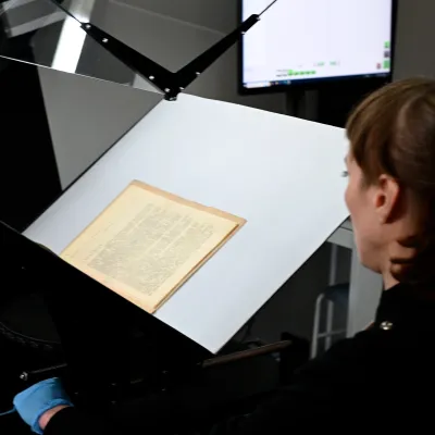 Osoba digitalizująca stary dokument przy użyciu stanowiska do skanowania; pożółkłe kartki leżą na podświetlanej powierzchni, a nad nimi znajduje się uchwyt z kamerą i ekranem komputerowym w tle.