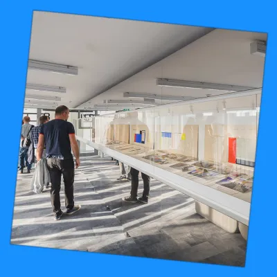 100 A photograph cropped to a crooked square. A group of people viewing an exhibition in a glass case.