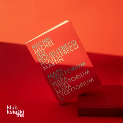 Na pomarańczowym tle stoi pod lekkim kątem książka Mapa i terytorium autorstwa Michel Houellebecq, oparta o niewielki czerwony podest. Czerwona okładka z powtarzającą się typografią imienia i nazwiska autora oraz bohatera przecina szeroki pas cienia, a w lewym dolnym rogu widnieje biały napis „klub książki ms”.