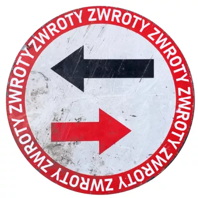 A circular road sign. The background of the sign is white, and a red circle runs around its perimeter. Inside this circle, on a white background, there are two horizontal arrows. The upper arrow is black and points to the left, while the lower arrow is red and points to the right. Along the red perimeter of the sign, in white letters, the word "TURNS" is repeated.
