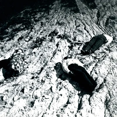 The black-and-white photograph depicts three limp human bodies lying on a steep, pale, and heavily undulating rocky slope. Two figures in dark trousers and light shirts, and one in a floral dress, rest motionless on their backs, creating a dramatic, unsettling composition.