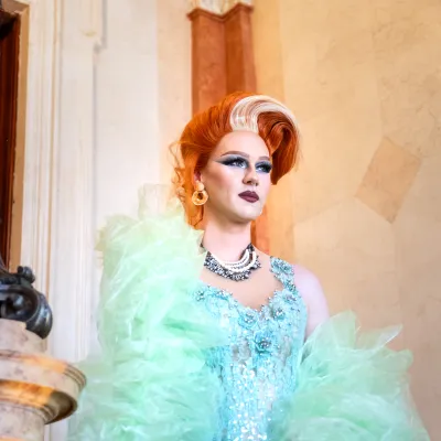 Portret drag queen z wysoką, rudą fryzurą i mocnym makijażem, ubranej w błyszczącą, błękitną suknię z puszystym, zielonym tiulem, pozującej w eleganckim, marmurowym wnętrzu.