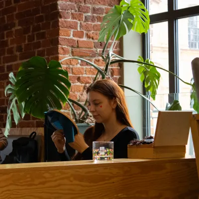 Młoda kobieta z rudymi włosami haftuje w pomieszczeniu z ceglaną ścianą, obok stoi duża monstera.