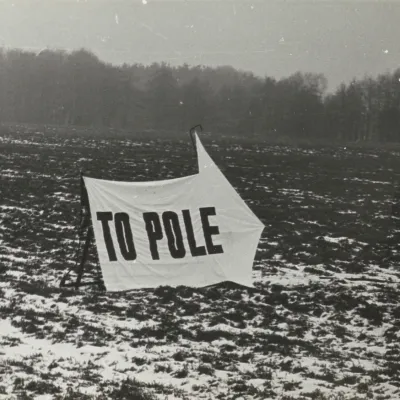 Czarno-białe zdjęcie przedstawia zaśnieżone pole, na którym ustawiono biały transparent z dużym, czarnym napisem „TO POLE”. Tkanina w kształcie strzałki, jest rozpięta na prostym stelażu i lekko powiewa na wietrze, a w oddali widać ciemną linię lasu.