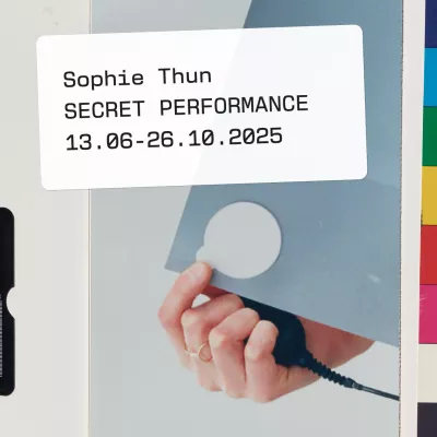 a hand holding something on the camera release cable, a black cable connected. The hand also holds a gray card with a white circle. The white frame in the center contains information about the exhibition. Sophie Thun. SECRET PERFORMANCE. From June 13 to October 26, 2025. On the right, a fragment of the color sampler